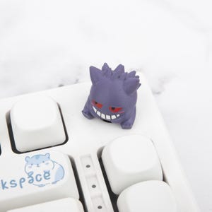 May include: A purple Gengar figure with red eyes and a wide grin rests on a white keyboard. The keyboard features white keycaps and a keycap with a blue hamster graphic and the word "kspace".