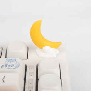 Peut inclure: Une figurine de lune en croissant jaune reposant sur une base de nuage blanc, fixée sur une touche de clavier mécanique blanche. Le clavier présente un graphique bleu clair d'un hamster avec le texte space imprimé sur une touche adjacente.