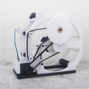 May include: A mechanical kinetic sculpture featuring a white frame with black rubber drive belts and gears. The device includes a central large white gear with a metallic bearing, smaller gears, and a blue wire connected to a white base component.