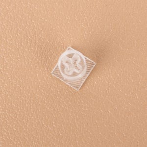 以下が含まれることがあります： 中央に十字型の開口部を持つ円形の部品を備えた、小型で透明な正方形の粘着パッド。透明なプラスチック製のベースには、小さなアイテムを固定するために設計された、格子状のテクスチャパターンが施されています。