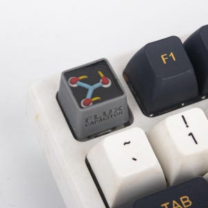 May include: A gray mechanical keyboard keycap featuring a black top surface with a light blue and red graphic of a flux capacitor. The side of the keycap is printed with the text FLUX CAPACITOR in a dark gray font, positioned on a white keyboard.