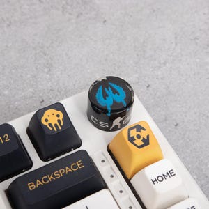 May include: A close-up of a mechanical keyboard with custom keycaps. The keycaps are black, yellow, and white. One keycap is a black cylinder with a blue and white design. Other keycaps have graphic designs. The keyboard is on a grey surface.