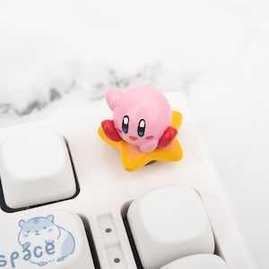 May include: A pink Kirby figurine with red arms and blue eyes, on a yellow star. The figurine is on a white keyboard, with a key featuring a blue hamster illustration and the word "space".