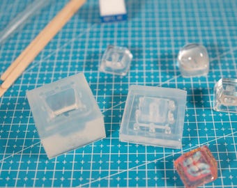 May include: A collection of translucent silicone molds for crafting mechanical keyboard keycaps, including a square base mold, a flat top mold, and several finished clear and pink resin keycaps, alongside wooden mixing sticks and a small blue box.