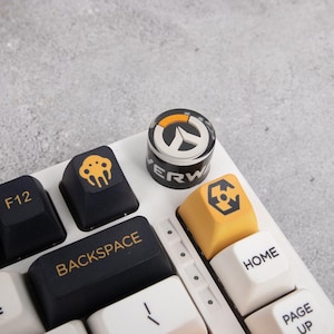 Op de afbeelding: Close-up van een wit mechanisch toetsenbord met aangepaste keycaps. De keycaps hebben verschillende ontwerpen en het woord "BACKSPACE". Eén keycap heeft het Overwatch-logo. Andere keycaps zijn zwart en geel.