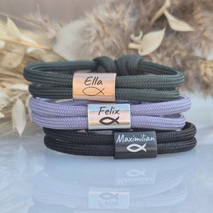 Könnte beinhalten: Drei personalisierte Armbänder mit den Namen "Ella", "Felix" und "Maximilian", die auf Metallplatten graviert sind. Jede Platte zeigt ein kleines Fischsymbol. Die Armbänder sind aus grünem, lila und schwarzem Seil gefertigt.