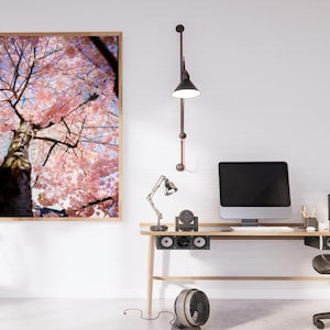 Puede incluir: Una obra de arte enmarcada representa un cerezo en flor con flores rosas y un tronco marrón. La obra cuelga sobre un escritorio de madera con un monitor de ordenador, una lámpara de escritorio y una silla de oficina negra. Una planta en maceta y una lámpara de pie también están en la habitación.