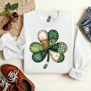 May include: White sweatshirt featuring a large, patchwork-style shamrock design in shades of green, tan, and white. The shamrock is composed of various fabric patterns and textures. The sweatshirt is a crew neck style.