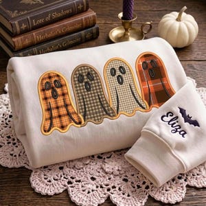 May include: A cream-colored sweatshirt featuring an embroidered design of four ghosts with various plaid patterns in orange, brown, and tan. The sleeve is embroidered with the name Eliza in dark purple script next to a small black bat silhouette.