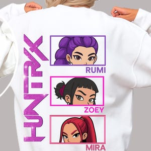 May include: White sweatshirt with a graphic design of three stylized female faces in rectangular frames. The names Rumi, Zoey, and Mira are below each face. The word "HUNTXK" is printed vertically on the left side in a glittery purple font.