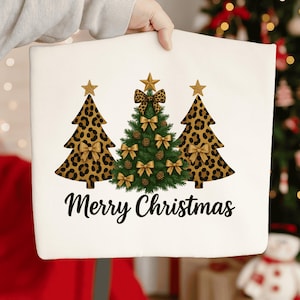 Könnte beinhalten: Weißes Kissenbezug mit Weihnachtsmotiv mit drei Weihnachtsbäumen. Zwei Bäume haben ein Leopardenmuster, und der mittlere Baum ist grün. Alle Bäume sind mit goldenen Schleifen und Sternaufsätzen verziert. Die Worte "Merry Christmas" sind in Schwarz geschrieben.