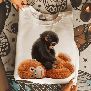 Könnte beinhalten: Ein weißes Sweatshirt mit einer detaillierten Illustration eines dunkelbraunen Affen, der auf einem orangefarbenen Plüschaffen sitzt. Der Plüschaffe hat ein lächelndes Gesicht. Das Sweatshirt ist auf einer gemusterten Oberfläche gefaltet.