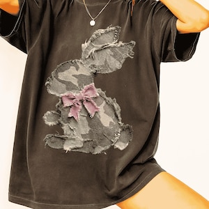 Pode incluir: T-shirt cinzenta escura com uma aplicação de coelho camuflado desgastado e um laço rosa. O design do coelho tem bordas desfiadas, adicionando uma estética vintage. A camisa tem um ajuste folgado e mangas curtas.