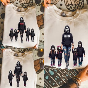 May include: A beige crewneck sweatshirt featuring a graphic print of a central figure wearing a ghost mask and a black hoodie with the text MAMA, surrounded by four smaller figures in matching ghost masks and black hoodies, some with pink bows.