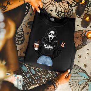 May include: Black sweatshirt featuring a graphic of a character in a Scream mask with long black hair, holding a coffee cup and making a peace sign. The word "MAMA" is printed on the sweatshirt. The sweatshirt is held by a person.