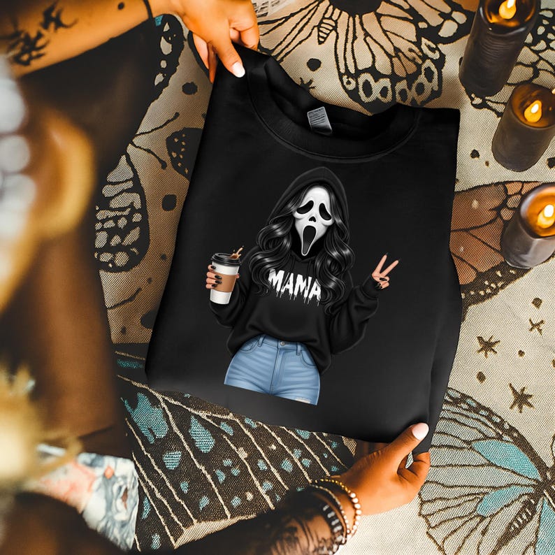 May include: Black sweatshirt featuring a graphic of a character in a Scream mask with long black hair, holding a coffee cup and making a peace sign. The word "MAMA" is printed on the sweatshirt. The sweatshirt is held by a person.