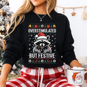 Może przedstawiać: Czarna bluza z szopem w czapce Świętego Mikołaja i łańcuchem światełek. Tekst "OVERSTIMULATED BUT FESTIVE" jest wyświetlany nad i pod grafiką szopa. Bluza ma świąteczny motyw.