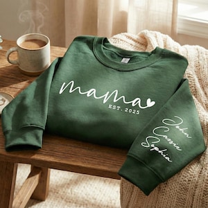 Könnte beinhalten: Ein dunkelgrünes Sweatshirt mit dem Wort "mama" in weißer Schrift, einem kleinen Herz und "EST. 2025". Der rechte Ärmel trägt die Namen "John", "Cassie" und "Sophia" in weißer Schrift. Eine Tasse Kaffee steht in der Nähe.