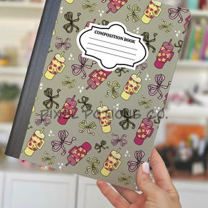May include: A composition book with a gray cover and a pattern of pink and yellow floral thermoses and bows. The label on the front reads "COMPOSITION BOOK". The notebook is held by a person.