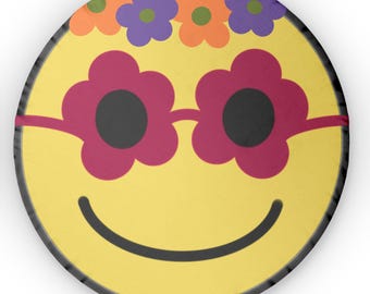 May include: A yellow smiley face with a black outline and a wide smile. The smiley face is wearing pink flower-shaped sunglasses and a crown of orange, purple, and green flowers.