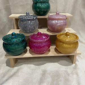 Puede incluir: Seis pequeños frascos redondos con tapa y purpurina incrustada en la resina. Los frascos son de varios colores, incluyendo rosa, verde, dorado y azul. Los frascos están dispuestos en un soporte de madera de dos niveles.