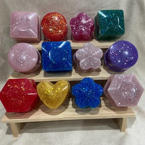 Könnte beinhalten: Ein Set aus 12 kleinen, dekorativen Boxen aus Harz mit eingearbeiteten Glitzer. Die Boxen haben verschiedene Formen, darunter quadratisch, rund, herzförmig, sternförmig und blütenförmig. Die Boxen sind auf einem dreistufigen Holzregal angeordnet.