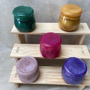 Op de afbeelding: Vijf kleine ronde potten met deksels, elk gevuld met een andere kleur glinsterende hars. De potten zijn gerangschikt op een houten, meertraps standaard. De kleuren zijn groen, goud, roze, paars en bruin.