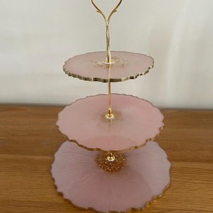 Può includere: Un vassoio a tre livelli con piatti in resina rosa e finiture dorate. I piatti hanno un bordo smerlato e dettagli glitterati dorati. Il supporto centrale e la maniglia sono dorati.