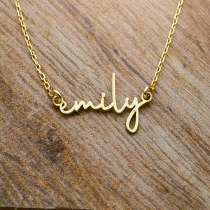 May include: A gold-toned necklace featuring a delicate chain and a central nameplate pendant displaying the name emily in a cursive script font. The metal has a polished finish and the pendant is securely attached to the chain on both sides.