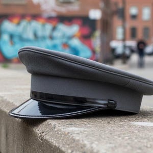 May include: A gray, peaked hat with a shiny black visor. The hat is resting on a concrete ledge. The hat has a structured shape and a dark band around the base of the crown.