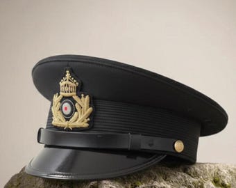 May include: A black military-style peaked cap featuring a stiffened visor and a black chin strap secured with a gold-toned button. The front displays an embroidered gold emblem with a crown, laurel leaves, and a central circular red and white target.