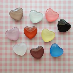 May include: Eleven heart-shaped glass beads in shades of brown, white, pink, red, yellow, and blue, arranged on a pink and white gingham background. The beads offer a range of colours and are ideal for craft projects or decorative purposes.