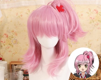 May include: A pink wig styled with a high ponytail and bangs, featuring a red cross-shaped hair accessory. The wig is displayed on a mannequin head. A small illustration of a character with a similar hairstyle is in the image.