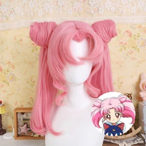 May include: A pink wig with a distinctive hairstyle featuring two high pigtails and long, flowing locks. The wig is displayed on a white mannequin head. A small cartoon graphic is also visible.
