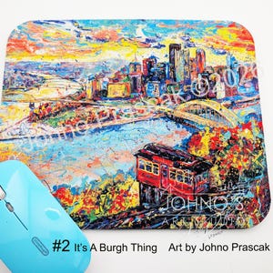 May include: A colorful mousepad featuring an abstract cityscape with a red cable car. The design includes a bridge, buildings, and water, with the text "#2 It's A Burgh Thing Art by Johno Prascak". A light blue wireless mouse is in the foreground.