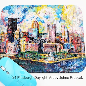 May include: A colorful mousepad featuring an abstract cityscape painting with buildings in shades of orange, purple, and blue. The mousepad has a blue border and the text "#4 Pittsburgh Daylight Art by Johno Prascak". A light blue wireless mouse is in the foreground.