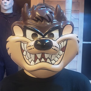 May include: A brown and tan plastic mask of the cartoon character Taz, the Tasmanian Devil, with a wide open mouth and sharp teeth. The mask has black eyes and a brown nose.