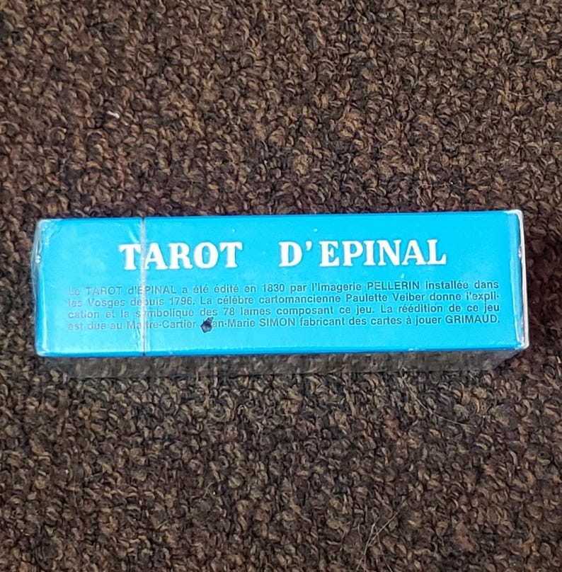 Puede incluir: Una caja azul con el texto "TAROT D'EPINAL" en blanco. La caja describe la baraja de tarot como editada en 1830 por la Imagerie Pellerin, establecida en los Vosgos desde 1796. Menciona a la famosa cartomanciana Paulette Velber que proporciona explicaciones y simbolismo para las 78 cartas. La reedici&oacute;n de esta baraja se debe a Jean-Mario Simon, fabricante de cartas de juego para Grimaud.