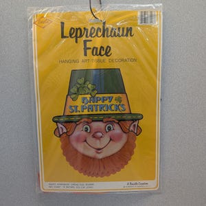 May include: A St. Patrick's Day hanging art-tissue decoration featuring a leprechaun face. The leprechaun has a red beard and rosy cheeks, wearing a green hat with shamrocks. The text "Happy St. Patricks" is displayed on the hat. The package is yellow.