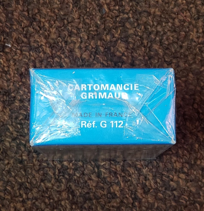 Puede incluir: Una caja azul con el texto "CARTOMANCIE GRIMAUD" y "R&eacute;f. G 112" impreso en ella. La caja est&aacute; envuelta en pl&aacute;stico.