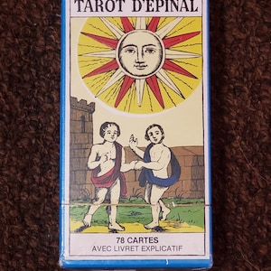 May include: A blue box with the text "Tarot d'Epinal" in white. The box features a yellow sun with a face and red rays. Two figures are depicted below the sun, standing in a green field. The text "78 Cartes" and "Avec Livret Explicatif" is printed on the bottom of the box.