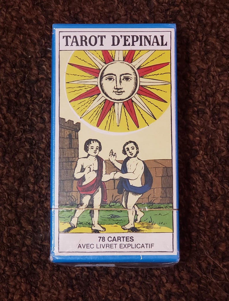 Puede incluir: Una caja azul con el texto "Tarot d'Epinal" en blanco. La caja presenta un sol amarillo con una cara y rayos rojos. Dos figuras se representan debajo del sol, de pie en un campo verde. El texto "78 Cartes" y "Avec Livret Explicatif" est&aacute; impreso en la parte inferior de la caja.