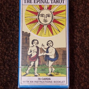 Puede incluir: Una caja azul con el texto "The Epinal Tarot" en letras blancas. La caja presenta una ilustraci&oacute;n de un sol con una cara y dos figuras que se toman de la mano. El texto "78 Cartas" y "Con un folleto de instrucciones" est&aacute; impreso en la parte inferior de la caja.