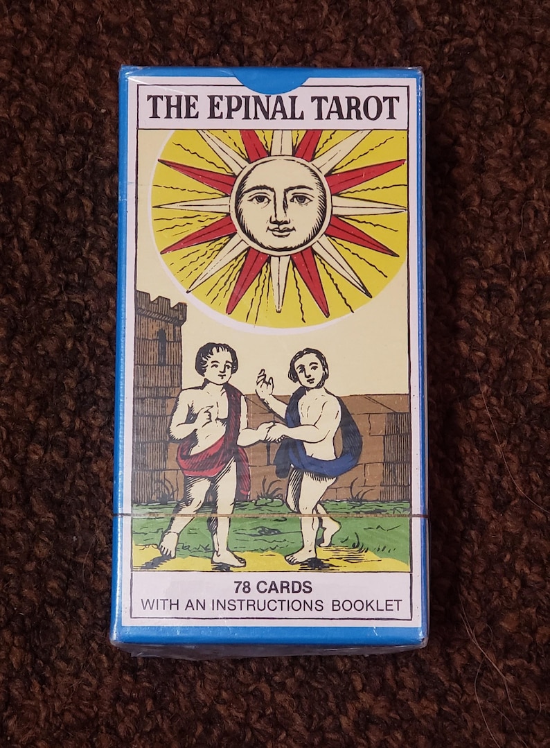 Puede incluir: Una caja azul con el texto "The Epinal Tarot" en letras blancas. La caja presenta una ilustraci&oacute;n de un sol con una cara y dos figuras que se toman de la mano. El texto "78 Cartas" y "Con un folleto de instrucciones" est&aacute; impreso en la parte inferior de la caja.