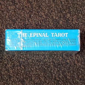Puede incluir: Una caja azul con el texto "The Epinal Tarot" impreso en ella. El texto explica que la baraja de tarot fue editada por primera vez en 1830 por impresores de arte llamados Pellerin, y que la baraja se ha impreso en Francia desde 1796.