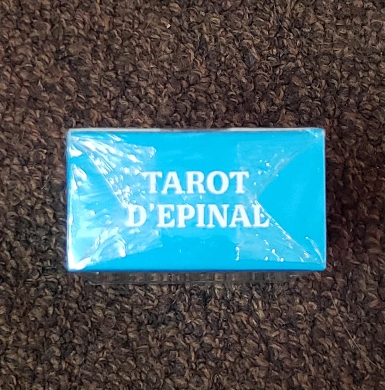 Puede incluir: Una caja azul con el texto "Tarot d'Epinal" impreso en blanco.