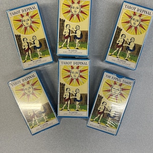 May include: Six decks of Tarot d'Epinal cards are arranged on a light gray surface. Each box features a yellow sun with a face, two figures, and the text "TAROT D'EPINAL". The boxes are blue with white text and illustrations.
