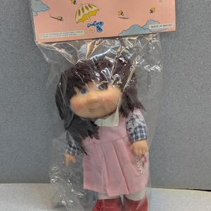 May include: A vintage Ann & Bob doll in a plastic package. The doll has dark hair, blue eyes, and is wearing a pink dress with a white collar and red shoes. The packaging includes the text "Ann & Bob" and illustrations of umbrellas.