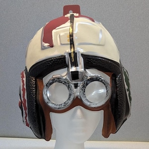 May include: A white and red pilot helmet with black trim and goggles. The helmet has a brown leather-like visor and ear coverings. The goggles are silver-rimmed with clear lenses. The helmet is displayed on a white mannequin head.