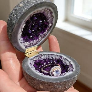 Op de afbeelding: Een geode ringdoos, opengeklapt om een sprankelend amethist interieur te onthullen. Een gouden en diamanten ring rust erin. De doos is opengehouden en toont de ring tegen de achtergrond van de paarse kristallen van de geode.